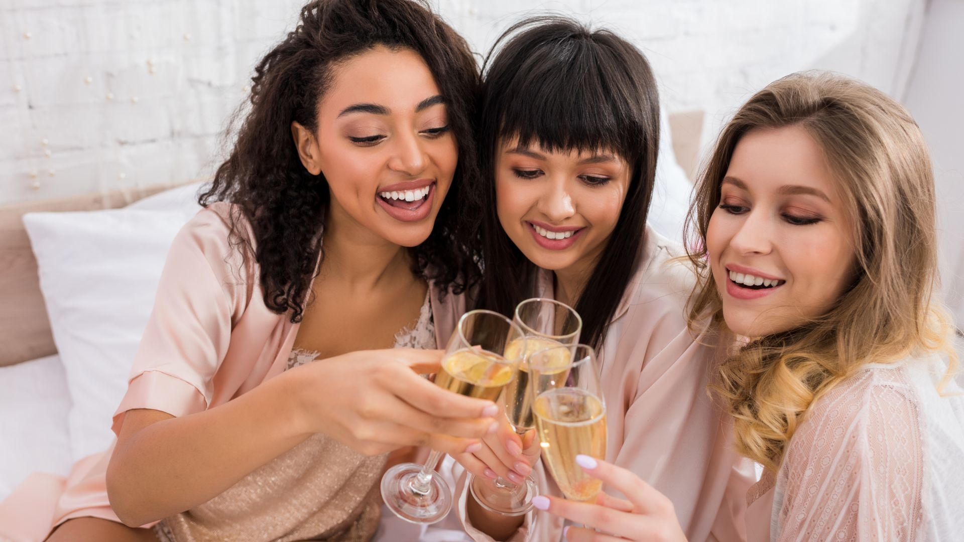 Three women toasting glasses of champagne on a bed