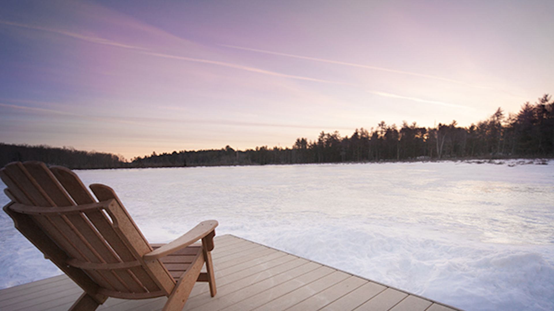 An Adirondack chair next to a frozen snow covered lake in Winter as the sun rises